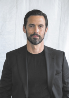 Fotografia de Milo Ventimiglia com expressão serena. Ele é branco, usa barba e tem cabelos curtos e lisos penteados para trás; está vestido com uma camiseta e um paletó.