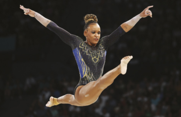 Fotografia de Rebeca Andrade. Ela realiza um salto na ginástica artística, com as pernas estendidas e os braços erguidos. Ela usa um collant preto com detalhes dourados e azuis. O cabelo está preso em um coque. Ao fundo, o público aparece desfocado.