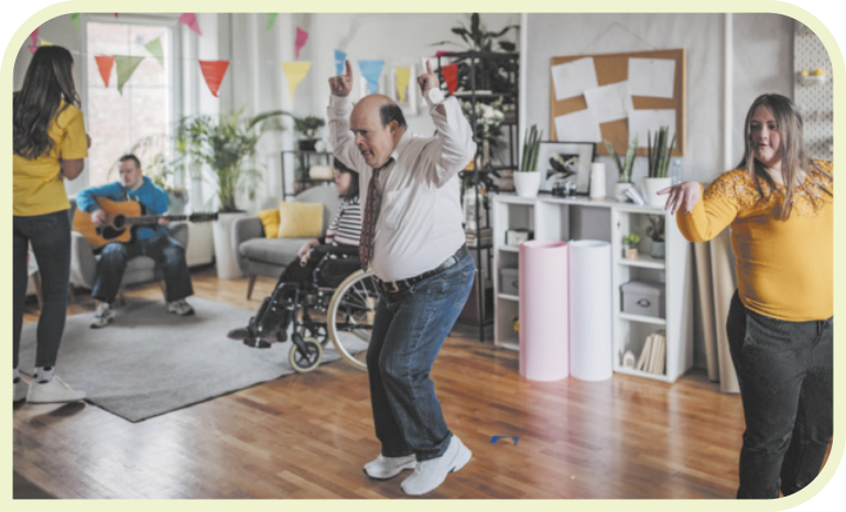 Fotografia de cinco pessoas dançando em uma sala-de-estar. Ao centro, há um homem calvo, usando camisa, gravata, calça jeans e tênis, com as mãos para cima e os joelhos flexionados. Ao lado dele, uma mulher loira movimenta as mãos. Ao fundo, vemos uma garota de costas, uma mulher em uma cadeira de rodas e um rapaz tocando violão, sentado em uma poltrona.