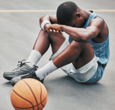 Fotografia de jogador de basquete sentado no chão de uma quadra, com os braços apoiados nos joelhos e a cabeça abaixada. Há uma bola de basquete em repouso chão, ao lado dele.