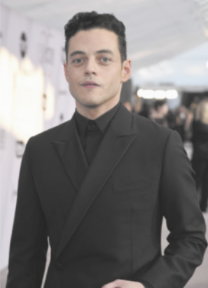 Fotografia de Rami Malek com expressão descontraída. Ele é branco, tem cabelos curtos e pretos e está usando camisa e paletó.