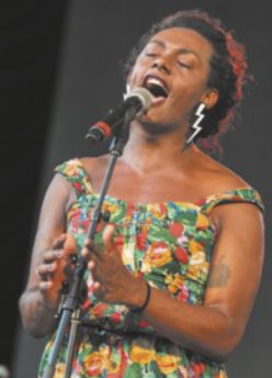 Fotografia de Liniker, cantando em um microfone de pedestal. Ela usa vestido estampado, cabelos curtos e brincos com formato de raio.