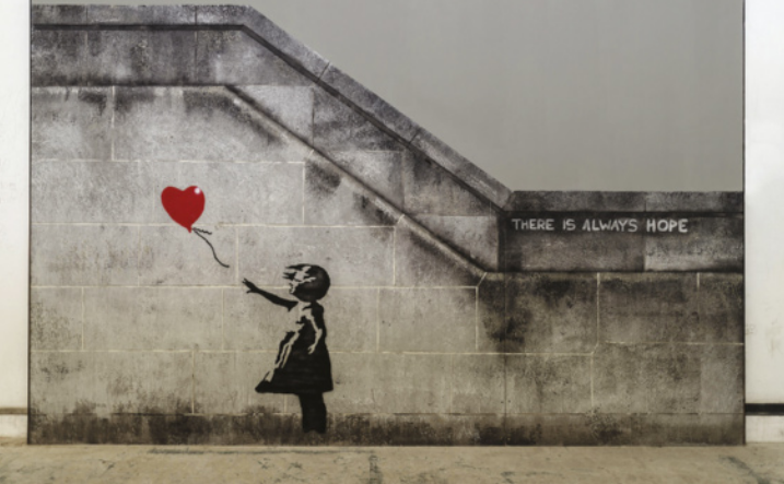Fotografia de um muro abaixo de uma escada, na rua, com um grafite. O grafite mostra a silhueta do perfil de uma menina soltando um balão vermelho em formato de coração, que flutua para longe. Os cabelos e o vestido da menina voam com o vento. Ao lado, na parede, está escrito: 'There is always hope.'