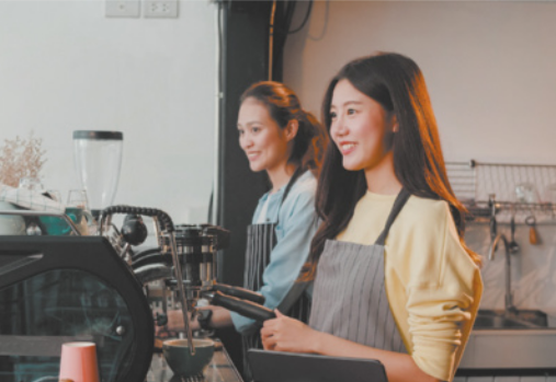 Fotografia de duas jovens trabalhando em uma cafeteria, sorrindo atrás do balcão, onde há diversos equipamentos como máquinas de café expresso. Elas têm traços asiáticos, cabelos longos e lisos e estão usando avental.