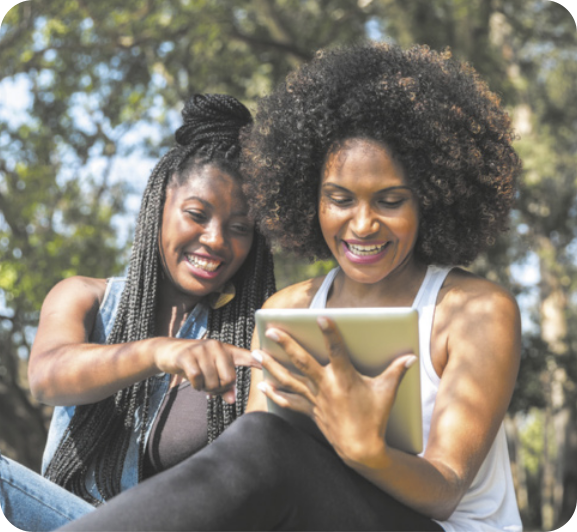 Fotografia de duas mulheres sorrindo, sentadas lado a lado. Uma segura um tablet, enquanto a outra aponta o dedo para a tela do aparelho. Ao fundo, há árvores e vegetação.