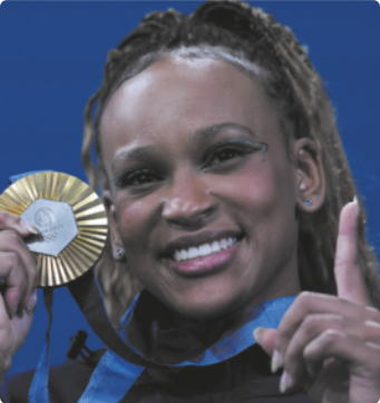 Fotografia de Rebeca Andrade sorrindo e exibindo uma medalha de ouro com uma das mãos, enquanto aponta para cima com a outra mão. Ela é negra e tem os cabelos trançados, presos para cima.