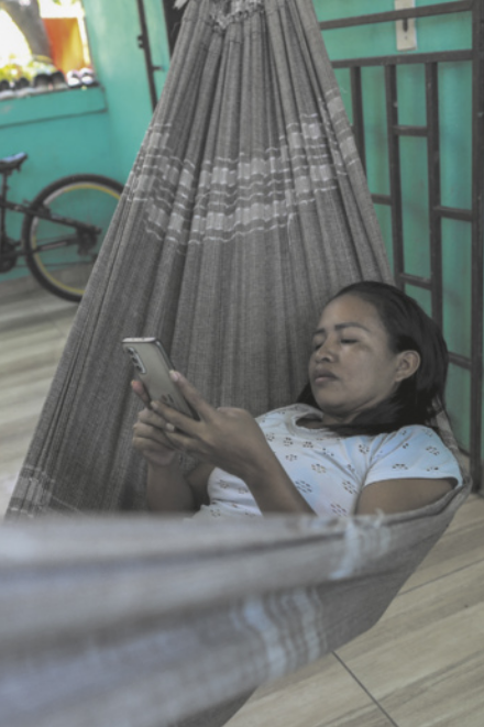 Fotografia de Cristina Quirino Mariano, uma mulher indígena, deitada em uma rede enquanto olha para a tela de um celular, fixamente. Ao fundo, há paredes verdes e uma bicicleta.