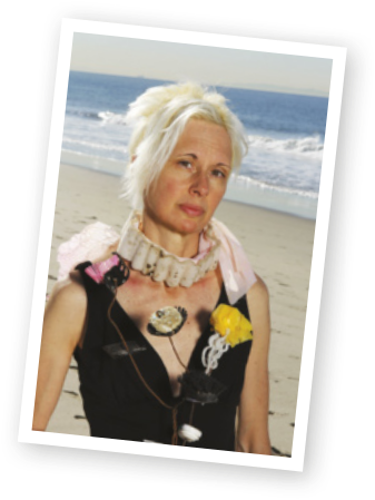 Fotografia de Marina DeBris, uma mulher branca, de cabelos curtos e brancos. Ela veste uma roupa preta, adornada com materiais descartáveis: uma embalagem plástica envolve seu pescoço e outros resíduos estão pendurados em sua roupa. Ao fundo, há uma praia.