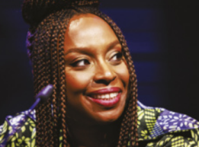 Fotografia de Chimamanda Adichie. Ela é uma mulher negra, com cabelos trançados, presos no alto da cabeça. Usa uma blusa estampada com tons de verde e azul e sorri levemente.