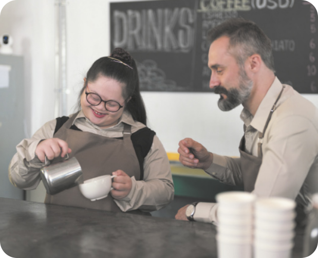 Fotografia de uma jovem preparando um café especial em uma cafeteria. Ela derrama controladamente o líquido de um bule de inox para uma xícara de porcelana. Ela é branca, tem traços de uma pessoa portadora da Síndrome de Down, usa óculos e está com os cabelos presos para trás. Ao seu lado, há um homem de barba e cabelo grisalhos observando. Ambos usam camisa e avental de uniforme da cafeteria.