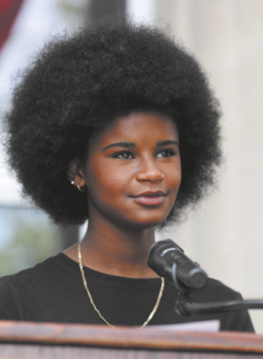 Fotografia de Marley Dias falando no microfone de um púlpito. Ela é negra, tem cabelos pretos no estilo Black Power e está usando colar no pescoço e blusa preta.
