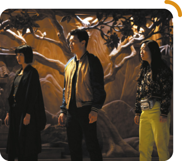 Cena do filme 'Shang-Chi and the Legend of the Ten Rings'. Na cena, três personagens, duas mulheres e um homem, estão parados, olhando para um mesmo lugar à frente deles. Todos têm traços asiáticos. Uma das mulheres tem cabelos curtos, pretos e lisos, e está usando sobretudo e calças; o homem tem cabelos curtos e pretos e está usando blusa de frio e calças; a outra mulher tem cabelos compridos e pretos e está usando camisa estampada, calças e uma pochete. Ao fundo, há uma pintura de rochas na parede e uma árvore.