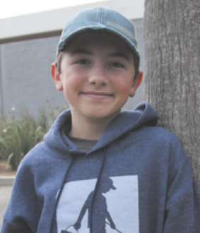 Fotografia de Ryan Hickman sorrindo, encostado no tronco de uma árvore. Ele é um menino branco, tem cabelos curtos e loiros e está usando boné e blusa de frio.