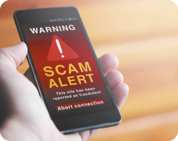 Fotografia de uma mão segurando um celular. Na tela do aparelho aparecem, em destaque, os avisos 'WARNING' e 'SCAM ALERT'. Entre os textos, há a imagem de um logotipo em forma de triângulo vermelho com um ponto de exclamação branco no centro, simbolizando alerta.