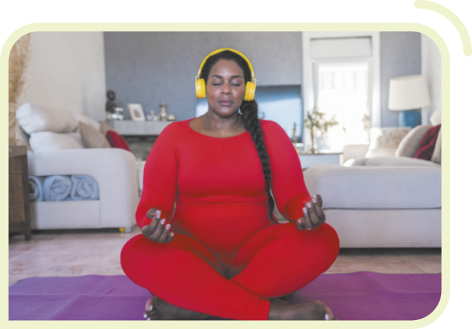 Fotografia de uma mulher meditando, sentada em um tapete em uma sala de estar. Ela está com as pernas cruzadas, o dorso das mãos apoiados nos joelhos, os olhos fechados e usando fones de ouvido. Ela é negra, tem cabelos longos com uma trança e está usando calça e blusa vermelhas.