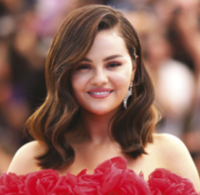 Fotografia de Selena Gomez. Mulher de pele branca e cabelos castanhos ondulados, penteados de lado e com comprimento até os ombros. Ela sorri levemente e usa um vestido vermelho. Ao fundo, há uma multidão desfocada.