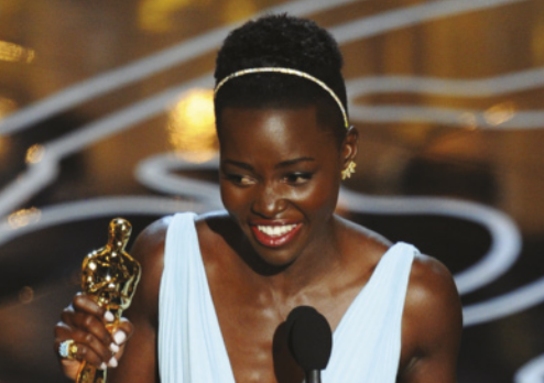 Foto de Lupita Nyong'o recebendo o prêmio Oscar de cinema. Ela está sorrindo com expressão emocionada atrás do púlpito, no palco, segurando a estatueta dourada. Lupita é negra, tem os cabelos curtos e crespos e está usando tiara e vestido branco decotado.