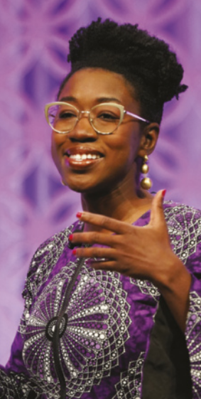 Fotografia de Joy Buolamwini. Ela é uma mulher negra, com cabelo curto e crespo, usa óculos e uma blusa estampada. Está sorrindo e faz um gesto com a mão, como se estivesse explicando algo.