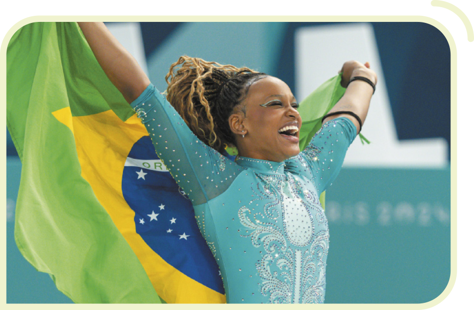 Fotografia da ginasta Rebeca Andrade sorrindo e segurando a bandeira do Brasil nas costas, com os braços abertos e esticados. Ela é negra, tem cabelos trançados, presos para cima, e está usando um collant com brilhos.