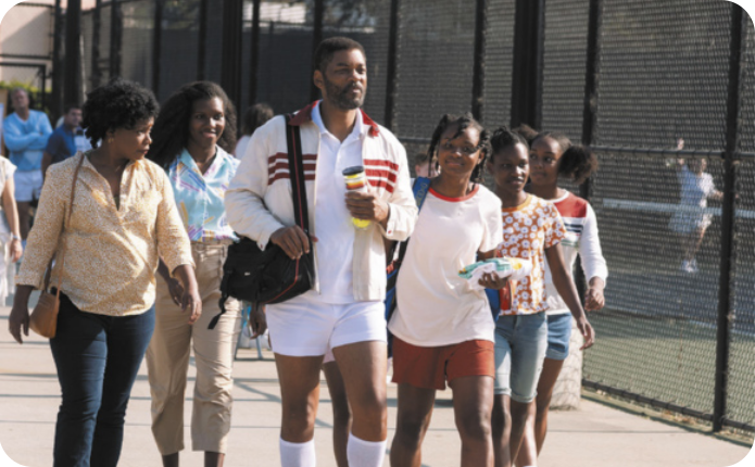 Cena do filme 'King Richard' mostrando o protagonista caminhando, acompanhado por duas mulheres e 3 meninas. Eles estão ao lado de uma quadra de tênis. Todos são negros. O protagonista está vestido com camiseta e shorts esportivos, um casaco e carrega uma bolsa e uma garrafa.