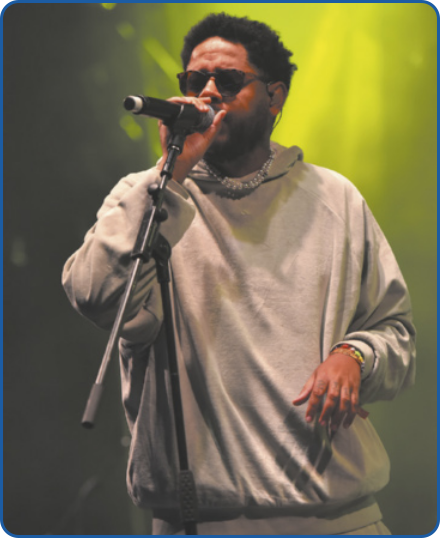 Fotografia de Emicida cantando ao microfone em um palco, durante um show. Ele é negro, tem cabelos curtos, pretos e crespos, e está usando óculos de sol, blusa de moletom, colar e pulseira.