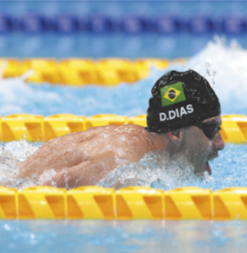 Fotografia de Daniel Dias na piscina. Ele tem pele branca e está usando óculos e touca de natação preta com a bandeira do Brasil e o nome 'D. Dias' estampado. Ele nada fazendo o estilo peito, com a cabeça fora da água e a boca aberta durante a respiração. Há raias delimitadoras amarelas na piscina.