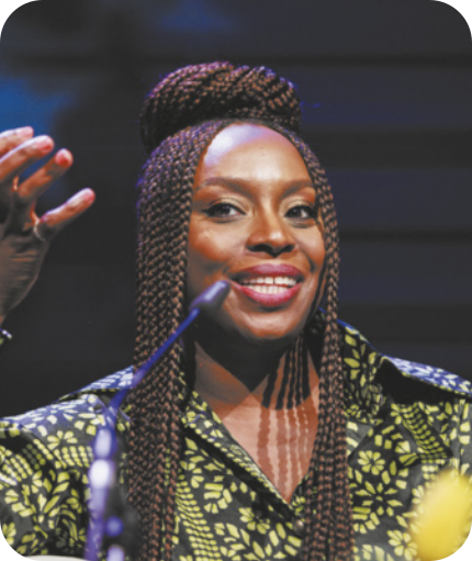 Fotografia de Chimamanda Ngozi Adichie. Ela é uma mulher negra, com tranças longas parcialmente presas em um coque alto no topo da cabeça. Veste uma blusa estampada colorida, está sorrindo e falando ao microfone, que está posicionado à sua frente.