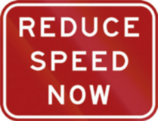 Imagem de uma placa de trânsito retangular com fundo vermelho e borda branca. No centro, está escrito em letras brancas: 'REDUCE SPEED NOW'.