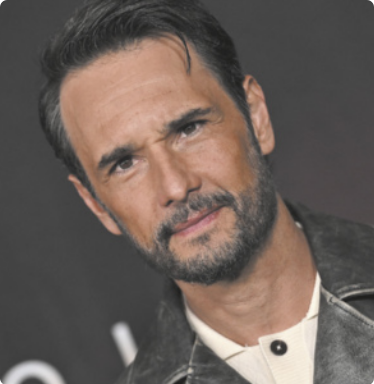 Fotografia de Rodrigo Santoro franzindo o cenho. Ele é branco, tem cabelos lisos, curtos, e barba e bigode grisalhos. Está vestido com uma camisa e uma jaqueta.