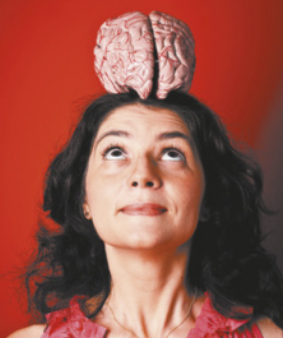 Fotografia de Suzana Herculano equilibrando um cérebro no topo de sua cabeça, olhando concentrada para ele. Ela é branca, tem cabelos ondulados e castanhos na altura dos ombros e está usando uma blusa vermelha.