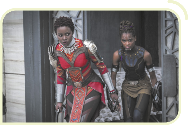 Cena de duas personagens do filme 'Pantera Negra' caminhando furtivamente. As atrizes são negras e usam figurino representando guerreiras. A personagem à frente tem cabelos curtos e cacheados. A outra personagem, atrás dela, tem cabelos longos com penteado de tranças.