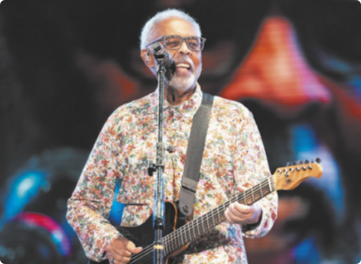 Fotografia de Gilberto Gil. Ele está em pé, em frente a um microfone de pedestal, segurando uma guitarra presa por uma alça ao ombro. Usa uma camisa estampada e tem os cabelos curtos, barba e bigode grisalhos.
