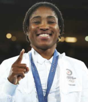 Fotografia de Cindy Ngamba sorrindo. Ela tem a pele negra e cabelos curtos e faz o gesto de número um com o dedo indicador levantado. Usa uma jaqueta branca com uma medalha pendurada no pescoço.