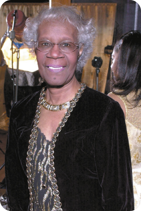 Fotografia de Shirley Chisholm. Ela está sorrindo; é uma mulher negra, com cabelos curtos, grisalhos e ondulados. Usa óculos, uma blusa preta e um colar.
