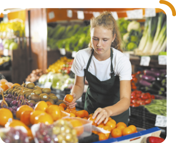 Fotografia de uma jovem organizando frutas na sessão de hortifrúti de uma mercearia. Ela é branca, tem cabelos loiros e longos, presos para trás, e está usando camiseta e avental.