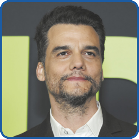Fotografia de Wagner Moura. Ele tem cabelos pretos, curtos e lisos, penteados para cima, barba e bigode. Veste uma camisa branca e um terno preto.
