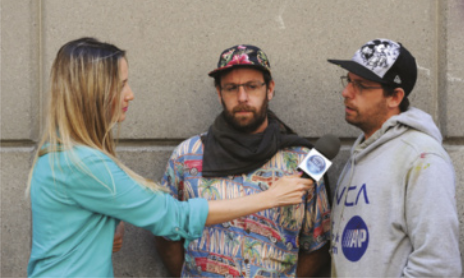 Fotografia dos irmãos gêmeos Otávio e Gustavo Pandolfo. Eles estão de pé, próximos a uma parede clara, sendo entrevistados por uma repórter. A repórter, com cabelos loiros e longos, usa uma blusa verde e segura um microfone. Otávio veste uma camiseta estampada e Gustavo, um moletom cinza com inscrições. Ambos usam bonés e óculos.