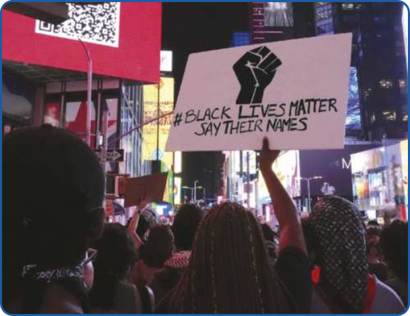 Fotografia de uma manifestação na rua. Diversas pessoas, a maioria negras, são vistas de costas. Uma mulher de cabelos trançados e compridos está levantando um cartaz com a imagem de um punho preto fechado e, abaixo, o texto: 'hashtag BLACK LIVES MATTER. SAY THEIR NAMES'.