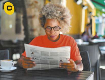 C. Fotografia de uma mulher sentada à mesa, lendo um jornal, com as mãos apoiadas sobre a superfície onde também há uma xícara. Ela tem cabelos grisalhos, ondulados e volumosos, usa óculos e uma camiseta vermelha. Ao fundo, há outras mesas e cadeiras.