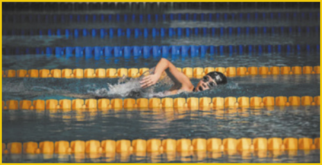 Fotografia de uma pessoa nadando em uma piscina. Ela está no centro da imagem, com o corpo parcialmente submerso, realizando o movimento de braçada. A piscina possui raias demarcadas por boias nas cores amarelo e azul.