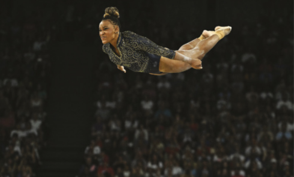 Fotografia da ginasta Rebeca Andrade durante um salto. Ela está em pleno ar, na horizontal, com o corpo esticado, os braços também esticados, ao lado do corpo, e olhando para frente com expressão de esforço. Ela usa collant preto com detalhes dourados e está com os cabelos presos em um coque.