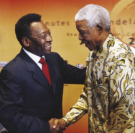 Fotografia de Pelé cumprimentando Nelson Mandela com um aperto de mãos. Pelé está à esquerda, é negro, tem cabelos curtos e crespos e está usando terno, camisa e gravata. Mandela está à direita, é negro, tem os cabelos curtos, brancos e crespos, e está usando uma blusa estampada de mangas longas. Ambos sorriem.