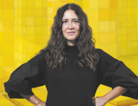 Fotografia de Adriana Varejão. Ele é uma mulher de pele branca e cabelos longos e ondulados, soltos sobre os ombros. Está em pé, com as mãos na cintura, usando uma blusa preta de mangas largas. O fundo é composto por tons de amarelo em padrão quadriculado.