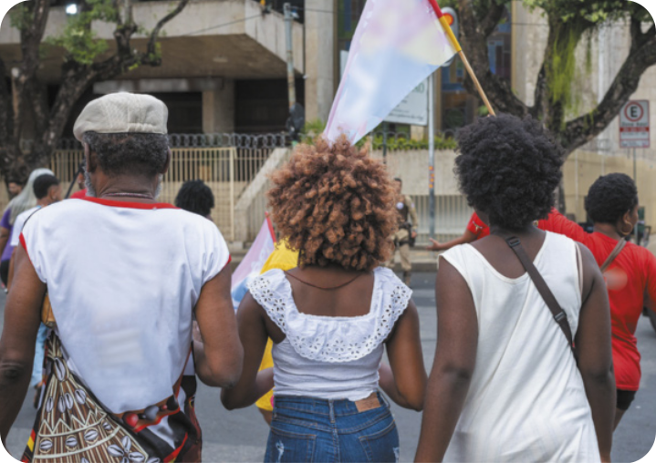 Fotografia de 3 pessoas negras caminhando lado a lado, vistas de costas. Há um homem de cabelos curtos, usando boina e camiseta sem mangas; uma mulher de cabelos loiros e volumosos, usando bata e jeans; e uma mulher de cabelos pretos e volumosos, usando vestido. Todos têm cabelos cacheados. À frente deles, uma quarta mulher negra carrega uma bandeira.