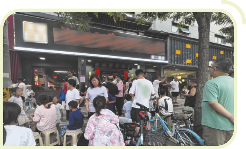 Fotografia de um grupo de pessoas reunidas em frente a um restaurante. Algumas estão sentadas em cadeiras plásticas, enquanto outras permanecem em pé, formando um aglomerado na calçada. Há bicicletas encostadas próximas à entrada do restaurante. Ao fundo, vê-se a fachada do prédio com letreiros e placas, além de algumas árvores.