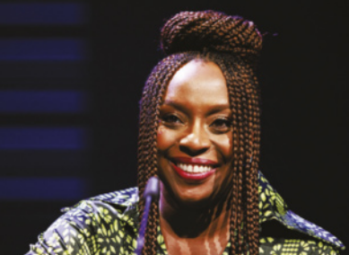 Fotografia de Chimamanda Ngozi Adichie sorrindo. Ela tem cabelo crespo longo, arrumado em tranças. Usa uma blusa estampada com cores e padrões variados.