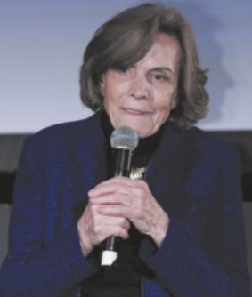 Fotografia de Sylvia Earle sentada, falando ao microfone, que segura com as duas mãos. Ela é idosa, branca, tem cabelos castanhos e lisos na altura da nuca e está usando uma blusa azul de mangas compridas.