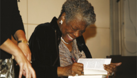 Fotografia de Maya Angelou. Ela é uma mulher idosa com cabelos curtos e grisalhos, sorrindo enquanto autografa um livro. Ela usa um casaco sobre uma blusa, colar e brincos.