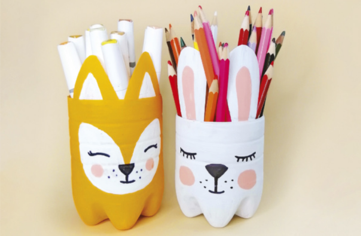 Fotografia de dois porta-lápis feitos com garrafas plásticas cortadas ao meio e pintadas com carinhas de animais. O da esquerda é amarelo com o desenho de uma raposa, contendo canetas brancas. O da direita é branco, com o desenho de um coelho, contendo lápis de cor. O fundo é bege claro.