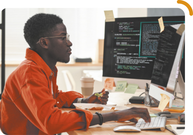 Fotografia de um jovem trabalhando em um computador, sentado à mesa. Ele digita com a mão direita enquanto segura um alimento com a mão esquerda. A embalagem com a comida está ao lado do teclado. Ele é negro, tem cabelos curtos e crespos, usa óculos e camisa de manga longa.